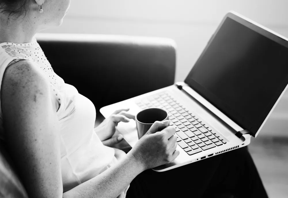 Kobieta pracująca na laptopie w fotelu, z kubkiem kawy w dłoni